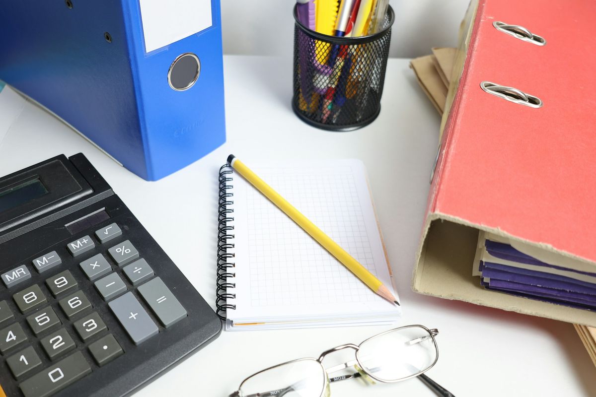 A calculator, a notebook with a pencil, reading glasses and two ring binders on a white desk