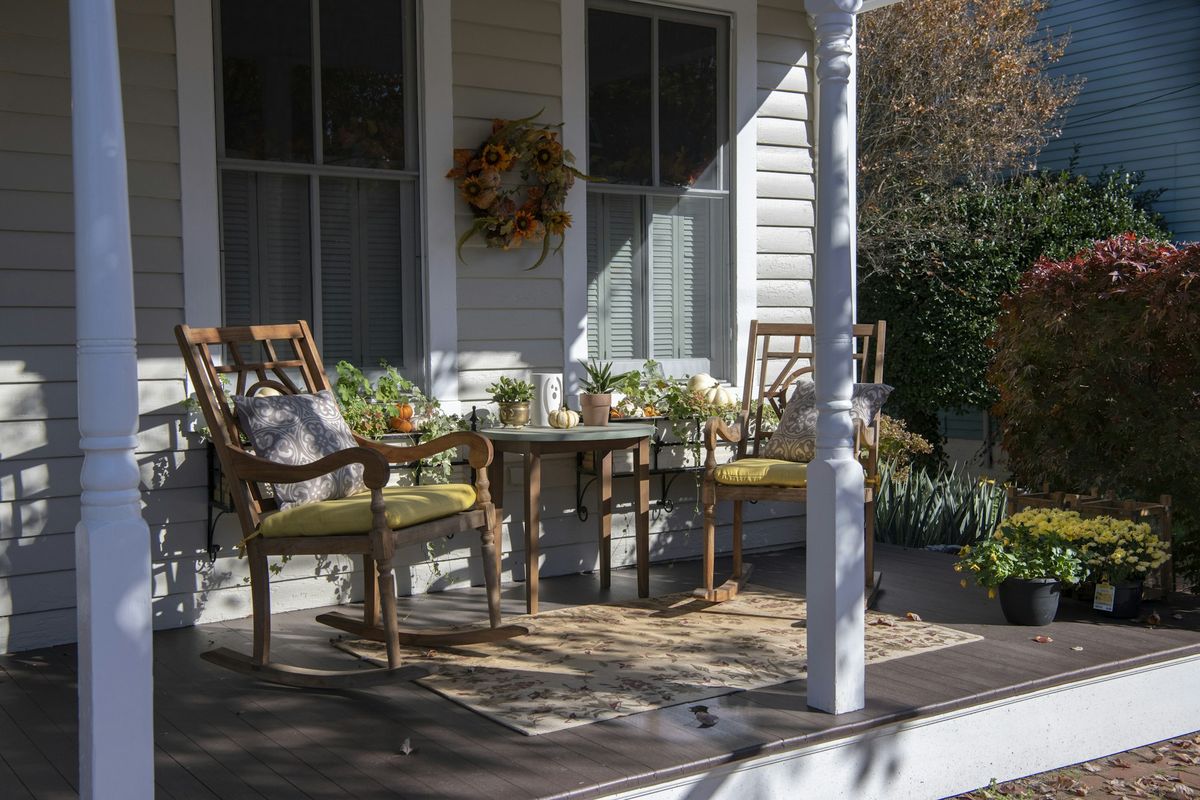 Front porch of an older white clapboard house with two wooden rocking chairs, yellow cushions, a small table and an autumn wreath