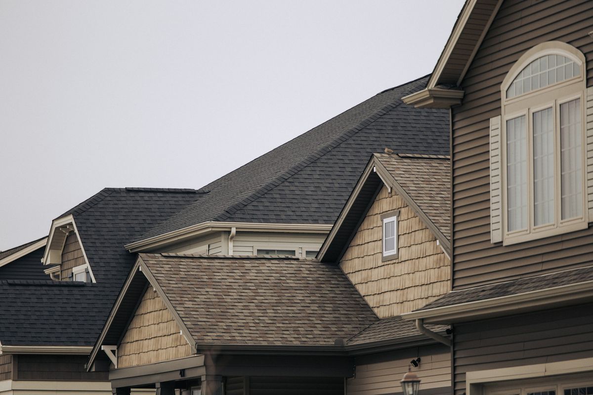 Rooftops of neighboring two-story suburban houses with gray and brown shingles, tan siding and white-trimmed windows under an overcast sky